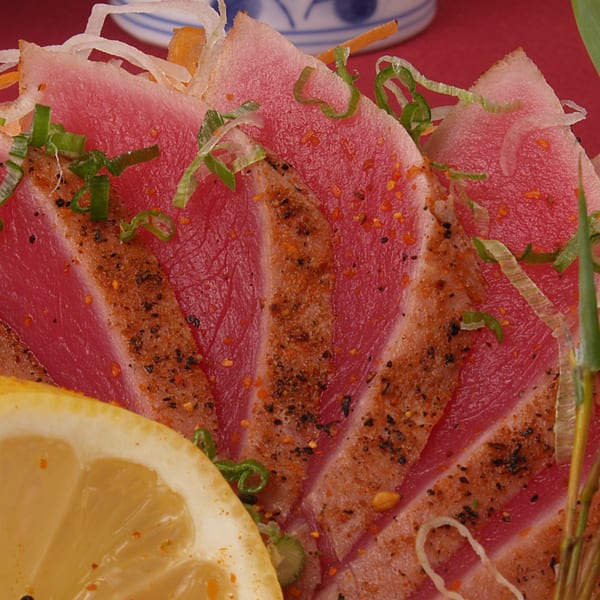Close-up of seared tuna sashimi slices, garnished with green onions, spices, and a lemon wedge.