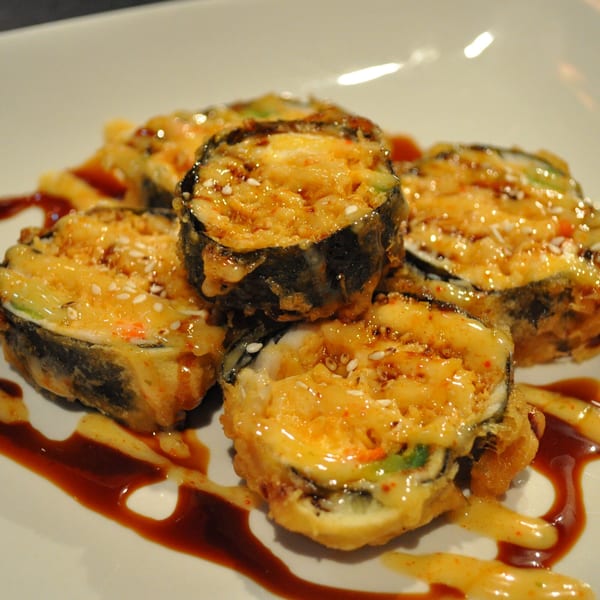 Deep-fried sushi rolls drizzled with sweet and spicy sauces, garnished with sesame seeds.