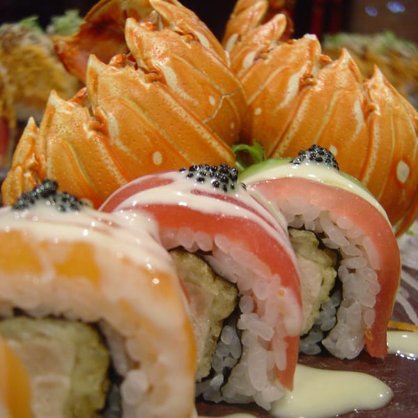 Sushi rolls topped with tuna, avocado, caviar, and sauce, with lobster tails in the background.