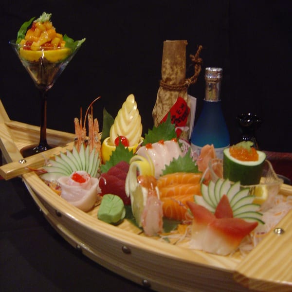 A wooden boat filled with various sushi and sashimi, with a cocktail and sake bottle in the background.