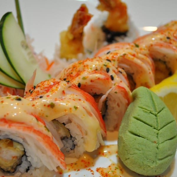 Sushi rolls topped with creamy sauce and spices, garnished with cucumber, lemon, and wasabi.
