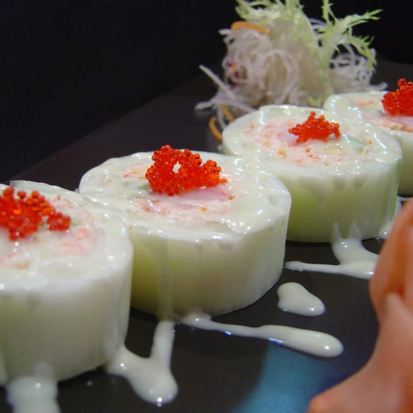 Four cucumber-wrapped sushi rolls topped with white sauce and bright red roe on a dark plate.